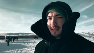 A person dressed in winter clothing, including a hooded jacket and beanie, stands in a snowy landscape. In the background, a child walks on the snow and a group of people stand near a building and a horse.