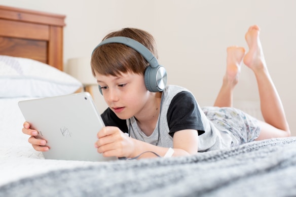 A child is lying on a bed with headphones on, intently using a tablet. The setting is a cozy bedroom with light-colored bedding and a wooden headboard.
