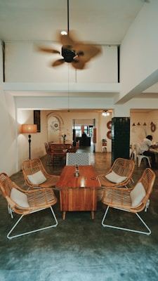 A cozy interior with rattan chairs arranged around a wooden coffee table on a polished concrete floor. A ceiling fan is spinning above, and the room leads into a dining area furnished with a variety of chairs and tables. The space is softly lit by a large standing lamp and complemented by wall decor. The overall ambiance is rustic with modern elements.