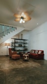 A cozy living room features a red sofa with patterned cushions, a matching armchair, and a small brown ottoman. A standing lamp is situated beside the armchair, casting a warm glow. An industrial-style bookshelf against the wall holds various decorative items. A staircase with a modern metal railing ascends in the background. The ceiling fan is in motion, creating a slight blur.