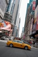 A bustling city street with tall skyscrapers and digital billboards. A yellow taxi is prominently featured, driving through an intersection. Pedestrians and vehicles crowd the urban scene, with vibrant advertisements displayed on buildings.