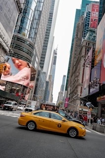 A bustling city street with tall skyscrapers and digital billboards. A yellow taxi is prominently featured, driving through an intersection. Pedestrians and vehicles crowd the urban scene, with vibrant advertisements displayed on buildings.