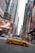 A bustling city street with tall skyscrapers and digital billboards. A yellow taxi is prominently featured, driving through an intersection. Pedestrians and vehicles crowd the urban scene, with vibrant advertisements displayed on buildings.