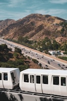 A white monorail train is passing in the foreground on an elevated track. Below, a busy highway with multiple lanes is visible, with numerous cars traveling in both directions. In the background, there are rolling hills covered in sparse vegetation under a partly cloudy sky.