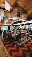A busy restaurant scene with a large group of people seated at a long table, engaging in conversation and enjoying their meals. Overhead, numerous light bulbs create a striking and modern chandelier design. The room features warm wooden flooring and artistic wall decorations with circular patterns.