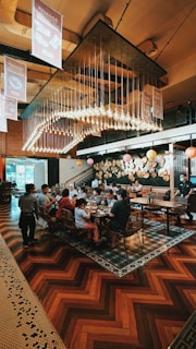 A busy restaurant scene with a large group of people seated at a long table, engaging in conversation and enjoying their meals. Overhead, numerous light bulbs create a striking and modern chandelier design. The room features warm wooden flooring and artistic wall decorations with circular patterns.