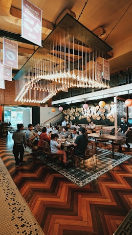A busy restaurant scene with a large group of people seated at a long table, engaging in conversation and enjoying their meals. Overhead, numerous light bulbs create a striking and modern chandelier design. The room features warm wooden flooring and artistic wall decorations with circular patterns.