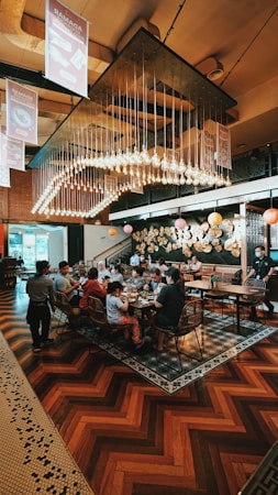 A busy restaurant scene with a large group of people seated at a long table, engaging in conversation and enjoying their meals. Overhead, numerous light bulbs create a striking and modern chandelier design. The room features warm wooden flooring and artistic wall decorations with circular patterns.