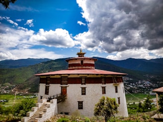 A traditional Bhutanese structure with a red roof and white stone walls is set against a backdrop of lush green hills and a dramatic sky filled with both fluffy white clouds and darker storm clouds.