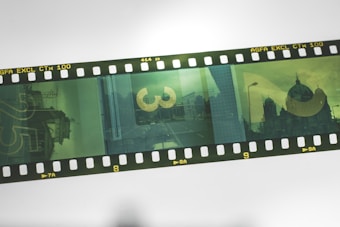 A strip of photographic film with multiple green-toned frames featuring various urban scenes, including a building with a dome and a street view. The film is labeled as AGFA EXCL CTx 100 and features both architectural elements and large numbers superimposed on the scenes.