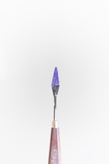 A small tool with a pointed, dark purple tip is centered against a plain light background. The handle is metallic, tapering down towards the tool's sharp end.