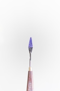 A small tool with a pointed, dark purple tip is centered against a plain light background. The handle is metallic, tapering down towards the tool's sharp end.