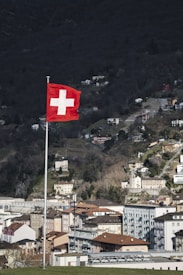 A Swiss flag prominently waves on a flagpole in the foreground. The background features a quaint European town nestled against a dark, forested hillside. The town showcases several pastel-colored buildings with brown roofs, surrounded by patches of greenery and small roads.