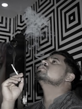 A man exhales smoke while holding a cigarette, set against a geometric black and white patterned wall. Smoke curls upwards in wisps above him. The lighting casts a dramatic effect, highlighting the patterns.
