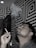 A man exhales smoke while holding a cigarette, set against a geometric black and white patterned wall. Smoke curls upwards in wisps above him. The lighting casts a dramatic effect, highlighting the patterns.