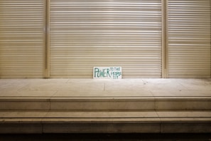 A handwritten sign with the words 'Power to the People' in green is propped against a closed, metal shutter in a seemingly deserted urban setting. The stark and minimalistic scene is emphasized by a series of steps leading up to the shutter, creating a sense of solitude and quietness.