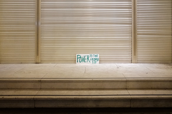 A handwritten sign with the words 'Power to the People' in green is propped against a closed, metal shutter in a seemingly deserted urban setting. The stark and minimalistic scene is emphasized by a series of steps leading up to the shutter, creating a sense of solitude and quietness.