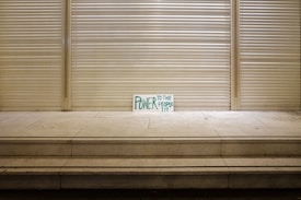 A handwritten sign with the words 'Power to the People' in green is propped against a closed, metal shutter in a seemingly deserted urban setting. The stark and minimalistic scene is emphasized by a series of steps leading up to the shutter, creating a sense of solitude and quietness.