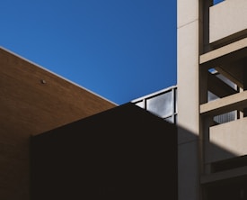 Geometric architectural forms are prominent, with angular shadows and lines. The composition includes a bright blue sky contrasted by the darker structures of a building, showcasing minimalist design elements.