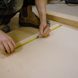 A person uses a measuring tape and pencil to mark measurements on a piece of light-colored wood. The individual is crouched down, wearing work boots and dark clothing, and is surrounded by a workbench setup.