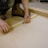 A person uses a measuring tape and pencil to mark measurements on a piece of light-colored wood. The individual is crouched down, wearing work boots and dark clothing, and is surrounded by a workbench setup.