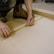 A person uses a measuring tape and pencil to mark measurements on a piece of light-colored wood. The individual is crouched down, wearing work boots and dark clothing, and is surrounded by a workbench setup.