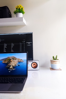 A clean and minimalistic workspace featuring a laptop displaying a nature-themed wallpaper, positioned on a glossy white desk. Behind the laptop is a larger monitor or TV screen showing a dark interface with text. A small speaker with an orange driver is situated next to the laptop. On the right, a small potted cactus sits in a decorative pot. Above these items, a shelf holds a white pot with a small green plant and a black bag.