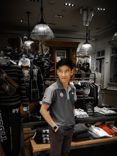 A man is standing inside a clothing store, surrounded by various garments on racks and shelves. The store is well-lit with modern ceiling lights and decorated with mannequins wearing casual outfits. Clothing items include shirts, jackets, and folded pants. There is a 'New Arrival' sign on display.