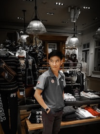 A man is standing inside a clothing store, surrounded by various garments on racks and shelves. The store is well-lit with modern ceiling lights and decorated with mannequins wearing casual outfits. Clothing items include shirts, jackets, and folded pants. There is a 'New Arrival' sign on display.