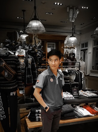 A man is standing inside a clothing store, surrounded by various garments on racks and shelves. The store is well-lit with modern ceiling lights and decorated with mannequins wearing casual outfits. Clothing items include shirts, jackets, and folded pants. There is a 'New Arrival' sign on display.