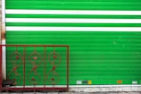 A green metal rolling shutter with two horizontal white stripes is partially visible. In front of it, there is a decorative red iron railing with intricate swirling patterns and floral motifs. The ground is concrete, and there are several small signs or stickers at the bottom of the shutter.