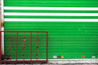 A green metal rolling shutter with two horizontal white stripes is partially visible. In front of it, there is a decorative red iron railing with intricate swirling patterns and floral motifs. The ground is concrete, and there are several small signs or stickers at the bottom of the shutter.