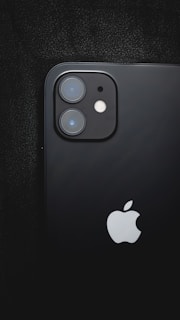 A close-up of a black smartphone with dual camera lenses and a flash on the back, featuring a prominent Apple logo in the lower section.