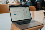 A MacBook Pro sits on a wooden table in a café setting. The screen displays a website advertising an easy way for Canadians to buy and sell bitcoin. A hand holding a smartphone is visible on the right side, and several people are sitting in the background, likely engaged in conversation.