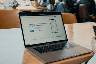 A MacBook Pro sits on a wooden table in a café setting. The screen displays a website advertising an easy way for Canadians to buy and sell bitcoin. A hand holding a smartphone is visible on the right side, and several people are sitting in the background, likely engaged in conversation.