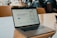 A MacBook Pro sits on a wooden table in a café setting. The screen displays a website advertising an easy way for Canadians to buy and sell bitcoin. A hand holding a smartphone is visible on the right side, and several people are sitting in the background, likely engaged in conversation.