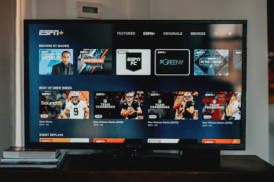 A television screen displaying the ESPN+ streaming interface with various sports-related shows and categories. The screen includes sections such as 'Browse by Shows' and 'Best of Drew Brees' with thumbnails of sports programs featuring athletes in action and analysts.