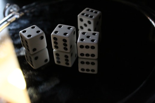 Four white dice with black dots are scattered on a reflective dark surface, with a partial view of a metallic object to the side. The dice display various numbers and are arranged randomly, casting reflections beneath them.