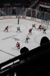 A hockey game is taking place in a large arena with players wearing red and white uniforms. The ice is marked with various lines typical of a hockey rink, and there are advertisements along the boards. A referee is on the ice, and a small crowd is visible in the stands. The players appear to be engaged in active play near one of the goals.