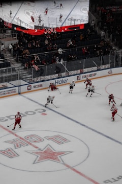 A hockey game is taking place on an ice rink inside an indoor arena. Players wearing red and white uniforms skate across the ice while engaged in the game. The stands are populated with spectators, watching the match attentively. Above the rink, a large electronic scoreboard displays a live view of the game.