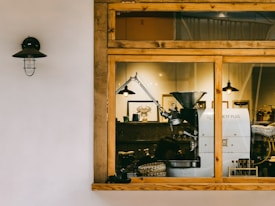 A cozy coffee shop interior is viewed through a large window. Inside, there is a coffee roasting machine prominently displayed, surrounded by wooden furnishings and warm lighting. The decor includes framed wall art and potted plants, contributing to a rustic and inviting atmosphere. The outside wall is simple and white with a black wall-mounted lantern.