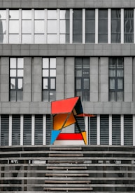 A modern art sculpture featuring geometric shapes in bright colors, including red, blue, yellow, and orange, positioned against the backdrop of a gray, industrial-style building with large windows. The sculpture contrasts sharply with the rigid lines and muted tones of the building behind it.