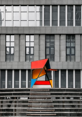 A modern art sculpture featuring geometric shapes in bright colors, including red, blue, yellow, and orange, positioned against the backdrop of a gray, industrial-style building with large windows. The sculpture contrasts sharply with the rigid lines and muted tones of the building behind it.