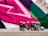 A collection of spray paint cans with various colored lids, including pink, yellow, and black, is arranged in front of a vibrant street art mural. The mural features bold geometric shapes in pink, green, and white, painted onto a textured wall. The ground is made of wooden planks, adding a natural contrast to the vivid colors of the artwork.