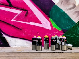 A collection of spray paint cans with various colored lids, including pink, yellow, and black, is arranged in front of a vibrant street art mural. The mural features bold geometric shapes in pink, green, and white, painted onto a textured wall. The ground is made of wooden planks, adding a natural contrast to the vivid colors of the artwork.