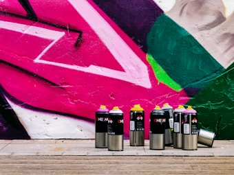 A collection of spray paint cans with various colored lids, including pink, yellow, and black, is arranged in front of a vibrant street art mural. The mural features bold geometric shapes in pink, green, and white, painted onto a textured wall. The ground is made of wooden planks, adding a natural contrast to the vivid colors of the artwork.