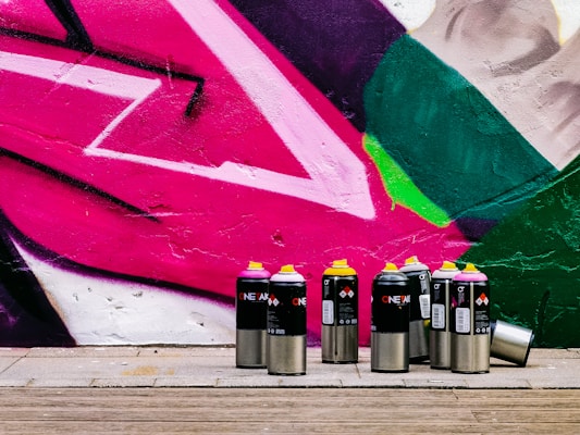 A collection of spray paint cans with various colored lids, including pink, yellow, and black, is arranged in front of a vibrant street art mural. The mural features bold geometric shapes in pink, green, and white, painted onto a textured wall. The ground is made of wooden planks, adding a natural contrast to the vivid colors of the artwork.