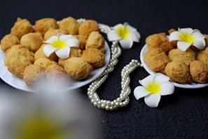 Two plates filled with golden-brown fried snacks, each adorned with a white and yellow frangipani flower. A pearl necklace is artistically draped around the plates, adding an elegant touch to the setup. The background is dark, highlighting the colors of the flowers and snacks.