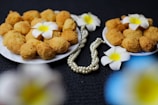 A close-up view of two plates filled with golden-brown fried snacks, garnished with white frangipani flowers. A strand of pearly beads is elegantly placed beside the plates on a dark textured background.