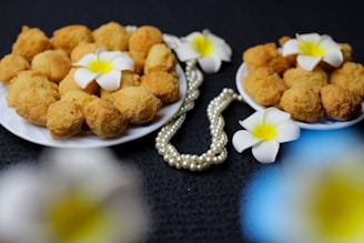 A close-up view of two plates filled with golden-brown fried snacks, garnished with white frangipani flowers. A strand of pearly beads is elegantly placed beside the plates on a dark textured background.